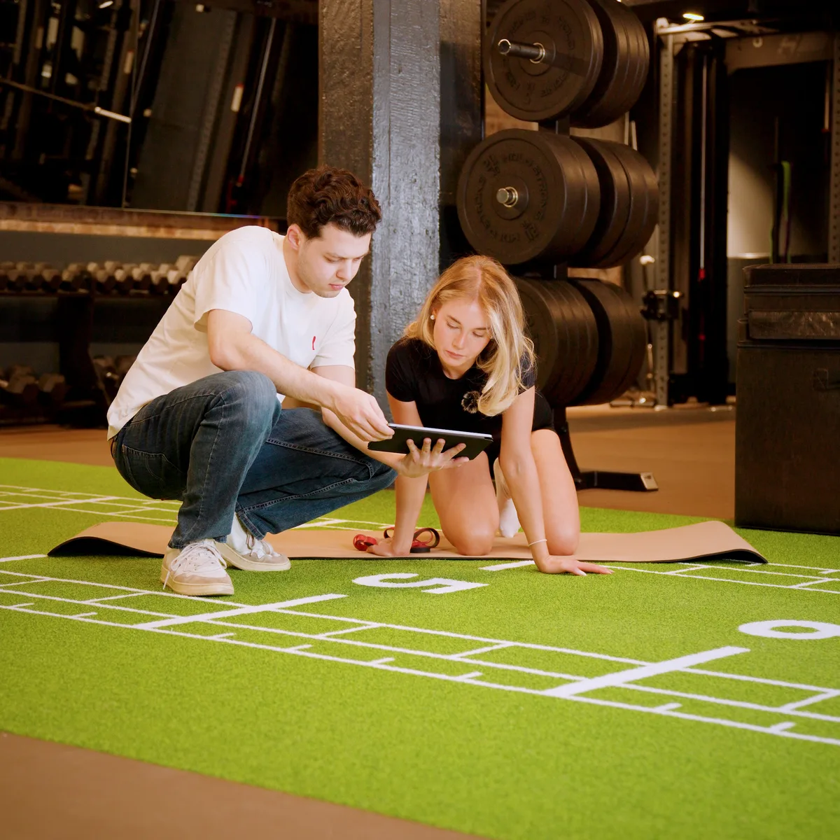 Coach und Sportlerin knien auf einer Trainingsmatte im Studio und schauen gemeinsam auf ein Tablet
